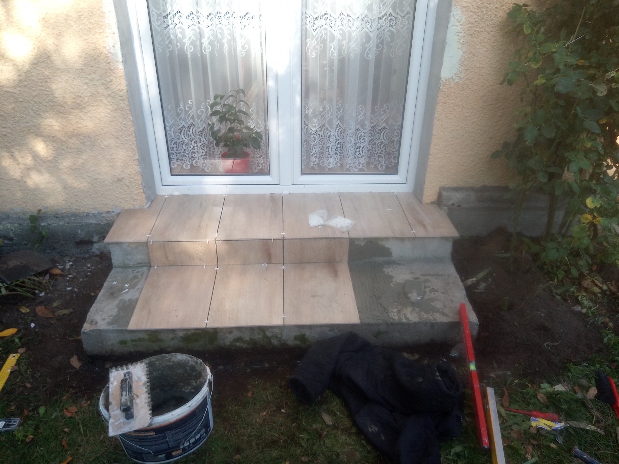 Układanie płytek na schodach wejściowych do domu, widoczny kubeł z zaprawą, szpachelka, poziomica i krzyżyki dystansowe.