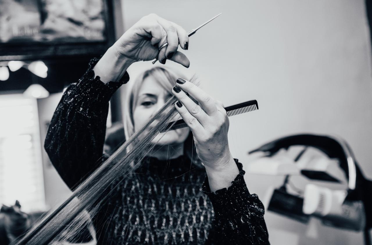 Czarno-białe zdjęcie kobiety w trakcie strzyżenia długich, jasnych włosów nożyczkami i grzebieniem, widoczne dłonie z ciemnym manicure, w tle sprzęt fryzjerski.