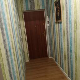 Sebastian Grabski - Korytarz po remoncie z drewnianą podłogą, pionowymi tapetami w odcieniach zieleni, błękitu i szarości, brązowymi drzwiami wejściowymi i obrazem w złotej ramie.