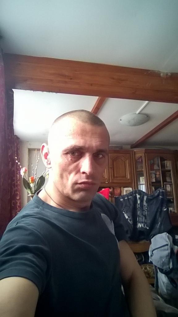 Mężczyzna z krótkimi włosami w pomieszczeniu z drewnianym sufitem i szafką w tle. Ujęcie z dołu.