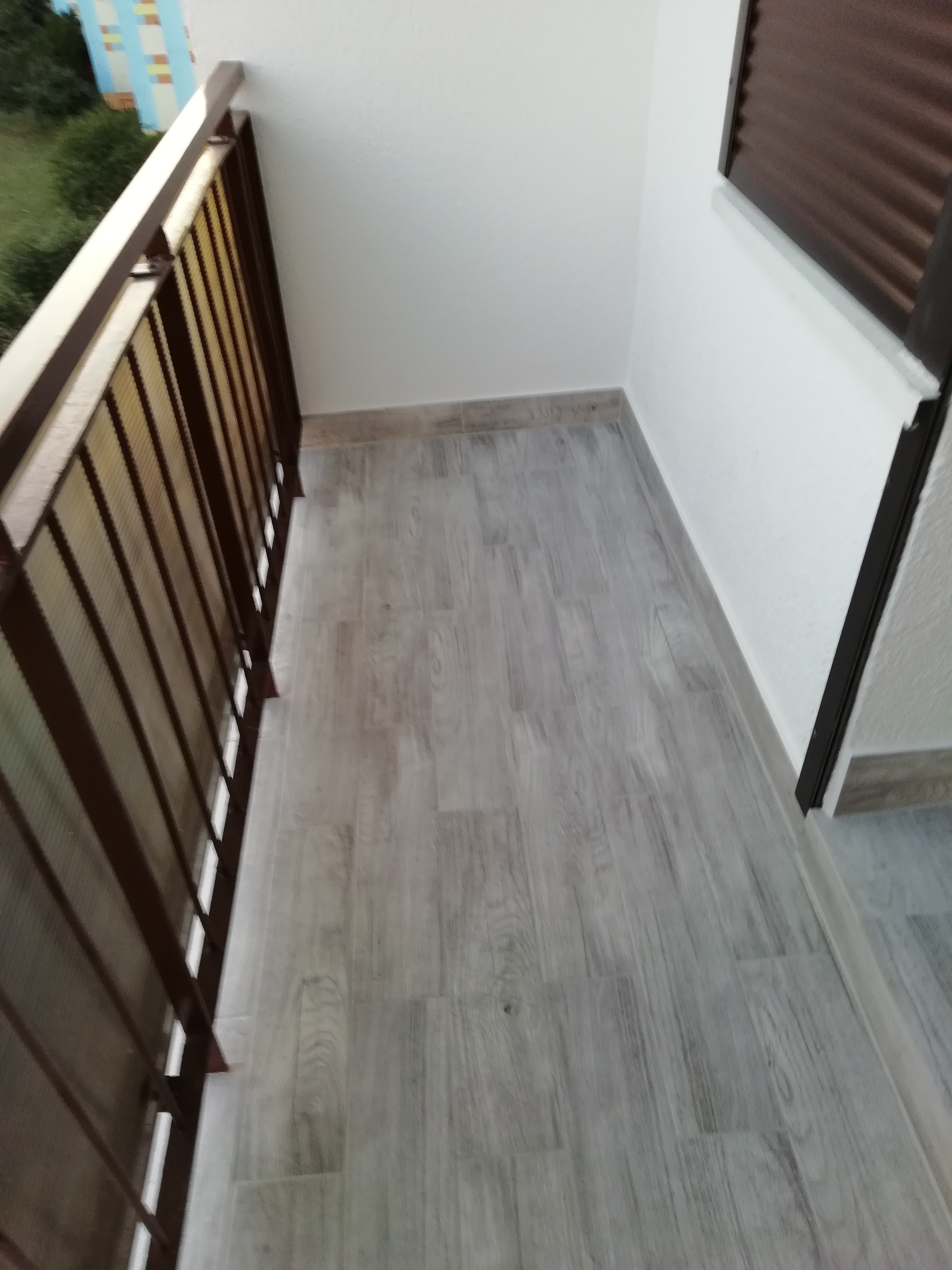 Wykończony balkon z szarą podłogą imitującą drewno, brązową metalową barierką i widocznym fragmentem okna z żaluzjami.
