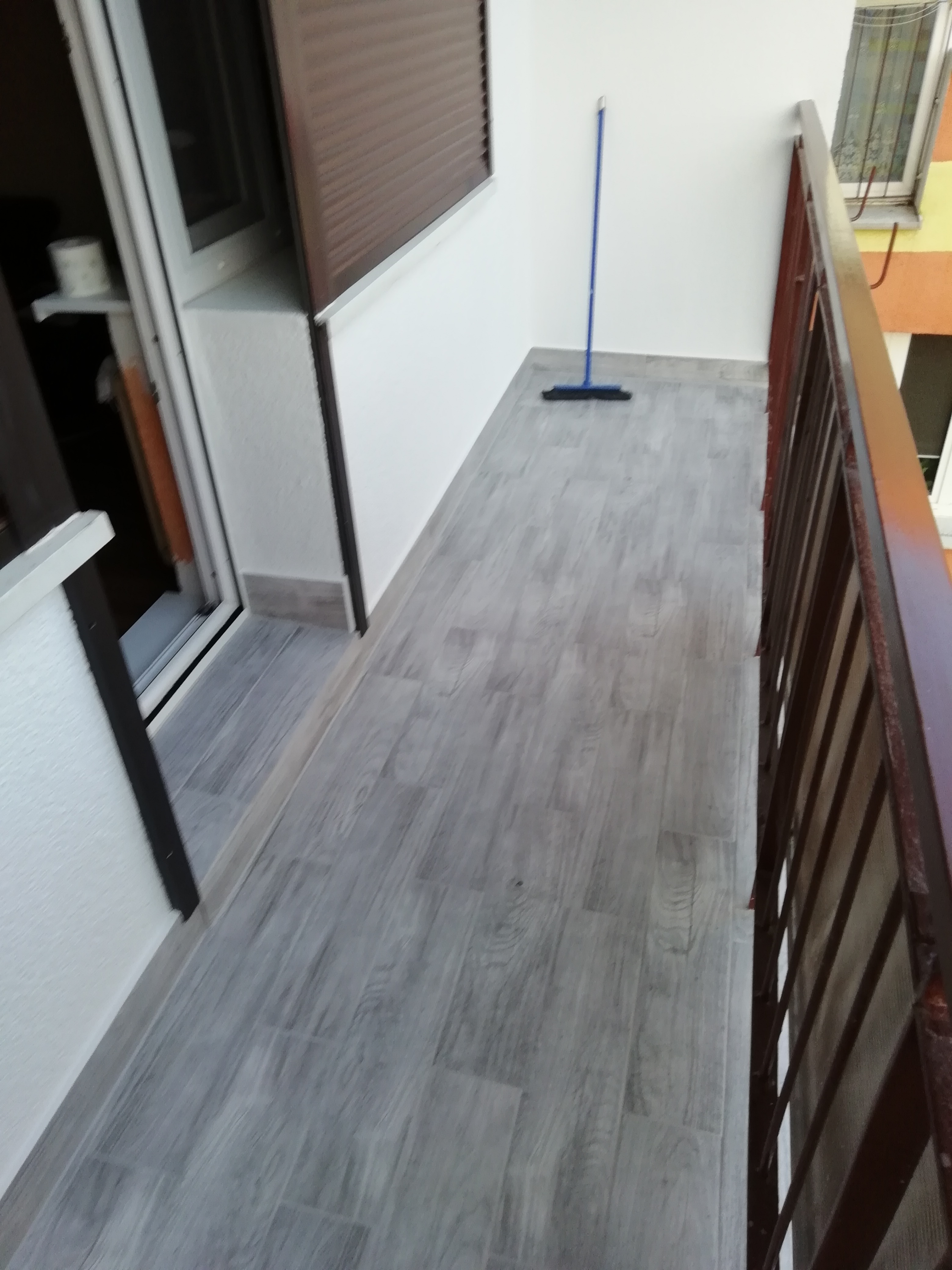 Wykończony balkon z szarymi płytkami imitującymi drewno, brązowa balustrada i niebieski mop w rogu, widok z góry.