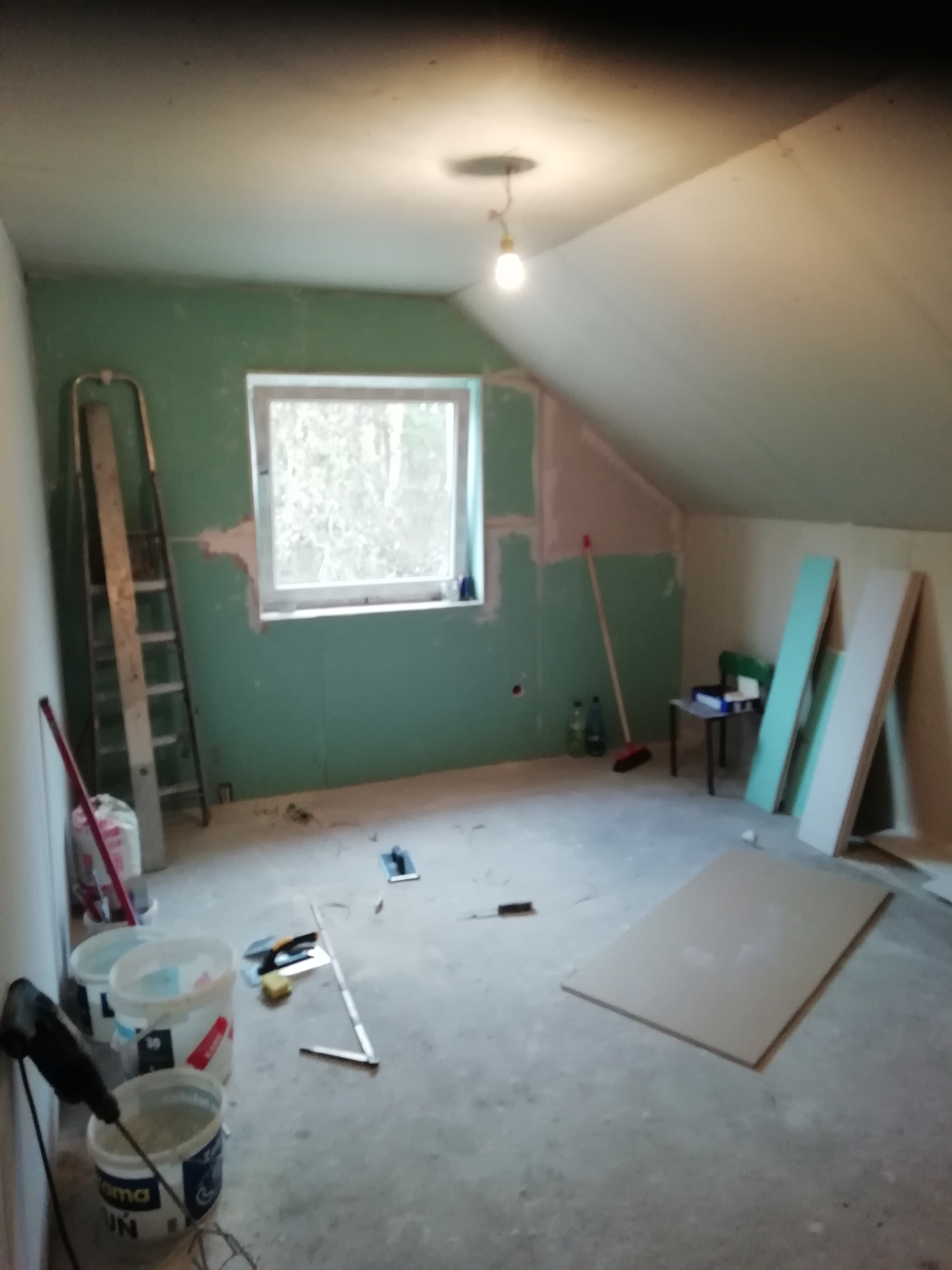 Poddasze w trakcie prac wykończeniowych: ściany częściowo pokryte zieloną farbą i gładzią, widoczne płyty kartonowo-gipsowe, drabina, wiaderka z zaprawą i narzędzia na podłodze.