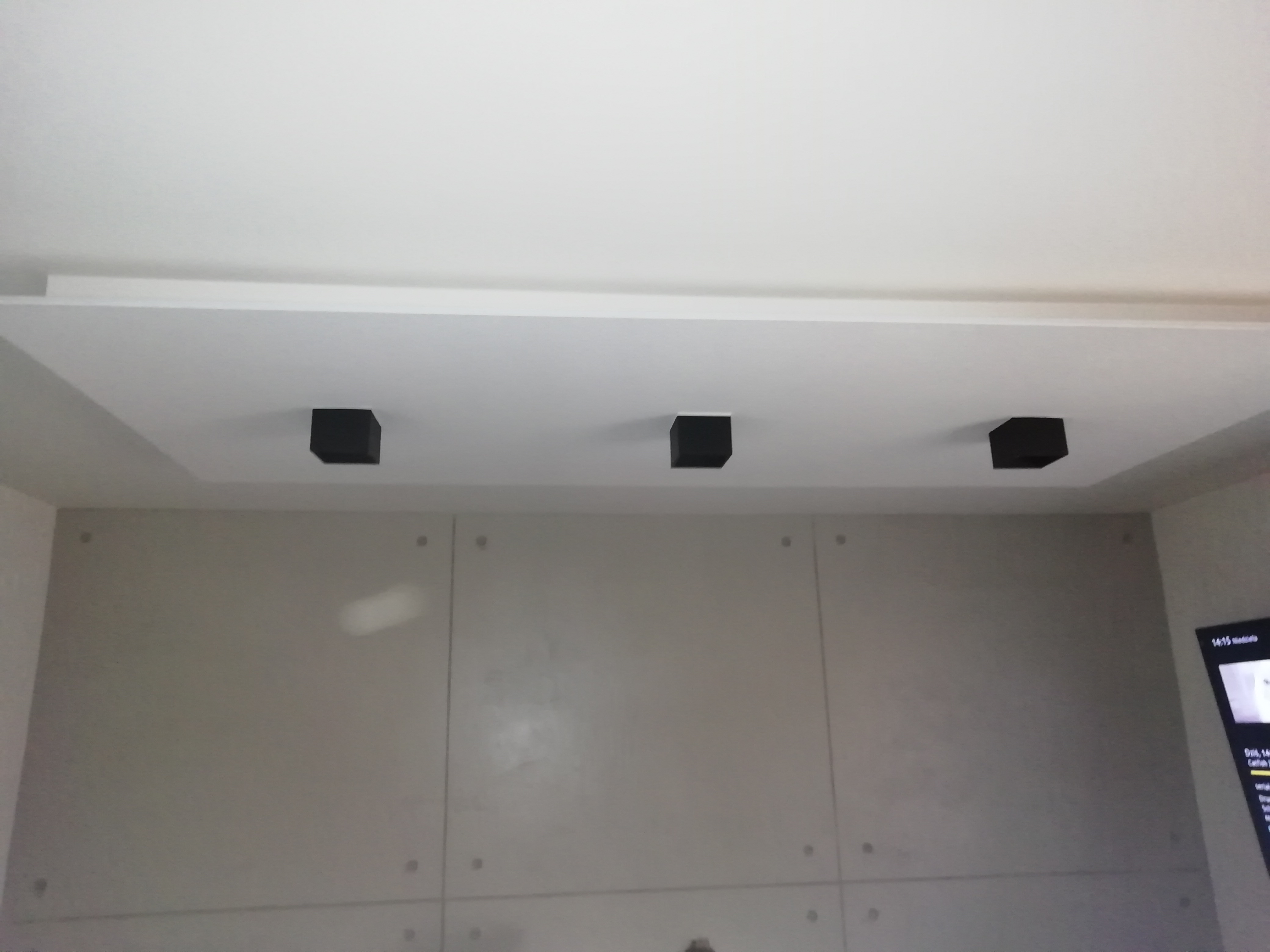 Minimalistyczne wnętrze: szara ściana z betonową fakturą, trzy czarne, kwadratowe lampy sufitowe wpuszczone w biały sufit podwieszany.