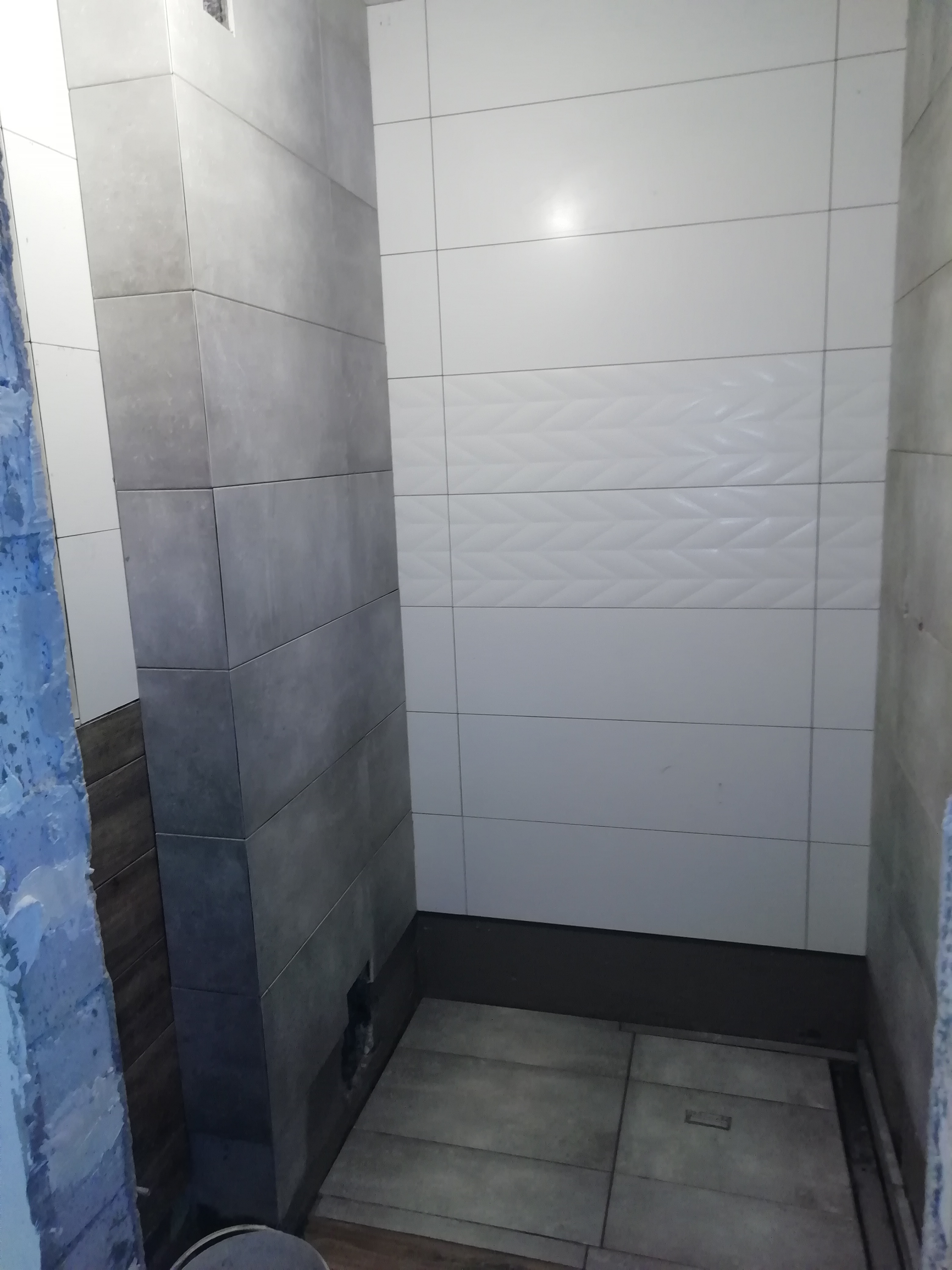 Świeżo wykafelkowana kabina prysznicowa z szarymi i białymi płytkami; białe płytki z subtelnym wzorem w jodełkę na ścianie; widoczne niedokończone krawędzie i ślady po taśmie malarskiej.