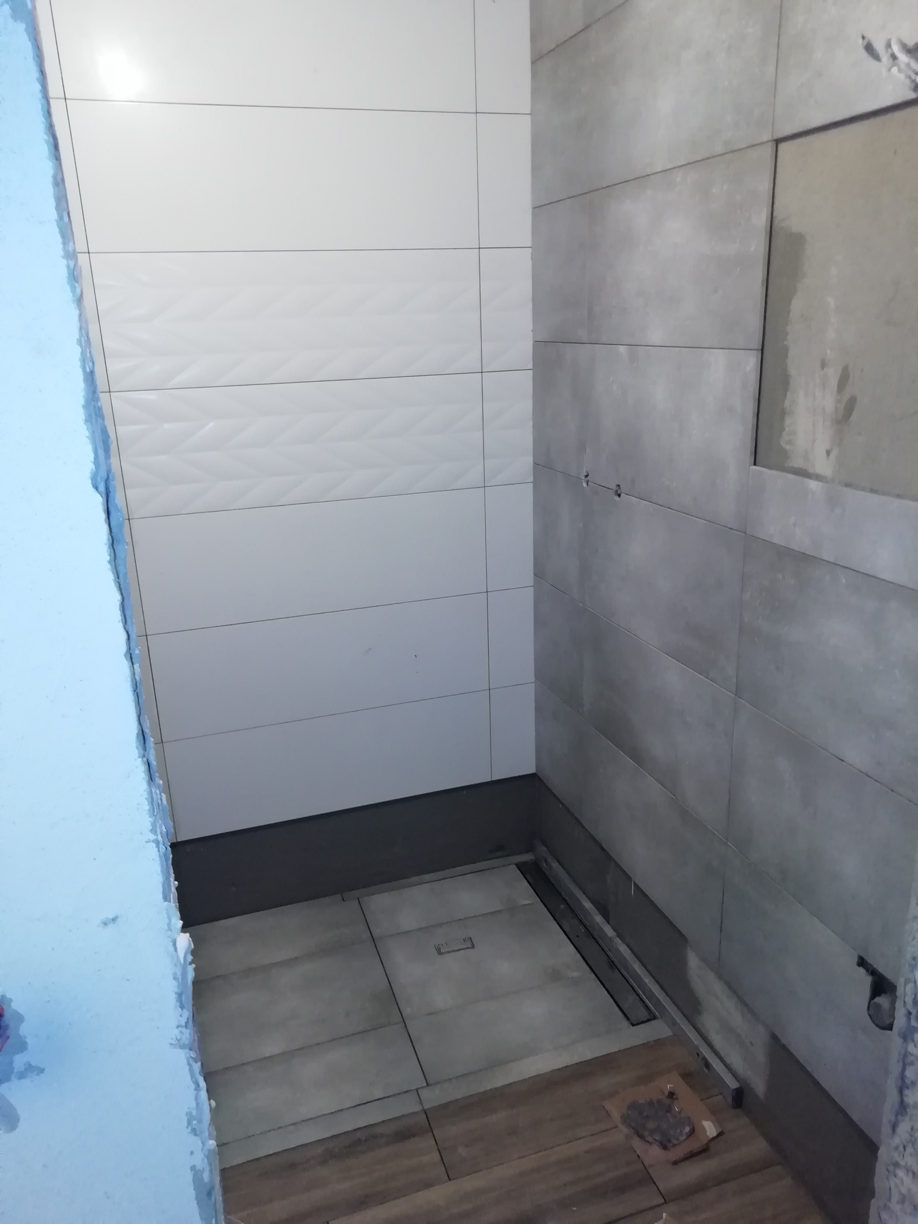 Wnętrze łazienki w trakcie wykańczania: białe płytki ścienne z subtelnym wzorem, szare płytki na sąsiedniej ścianie, widoczny odpływ liniowy w podłodze i fragment nieotynkowanej ściany z niebieskim...