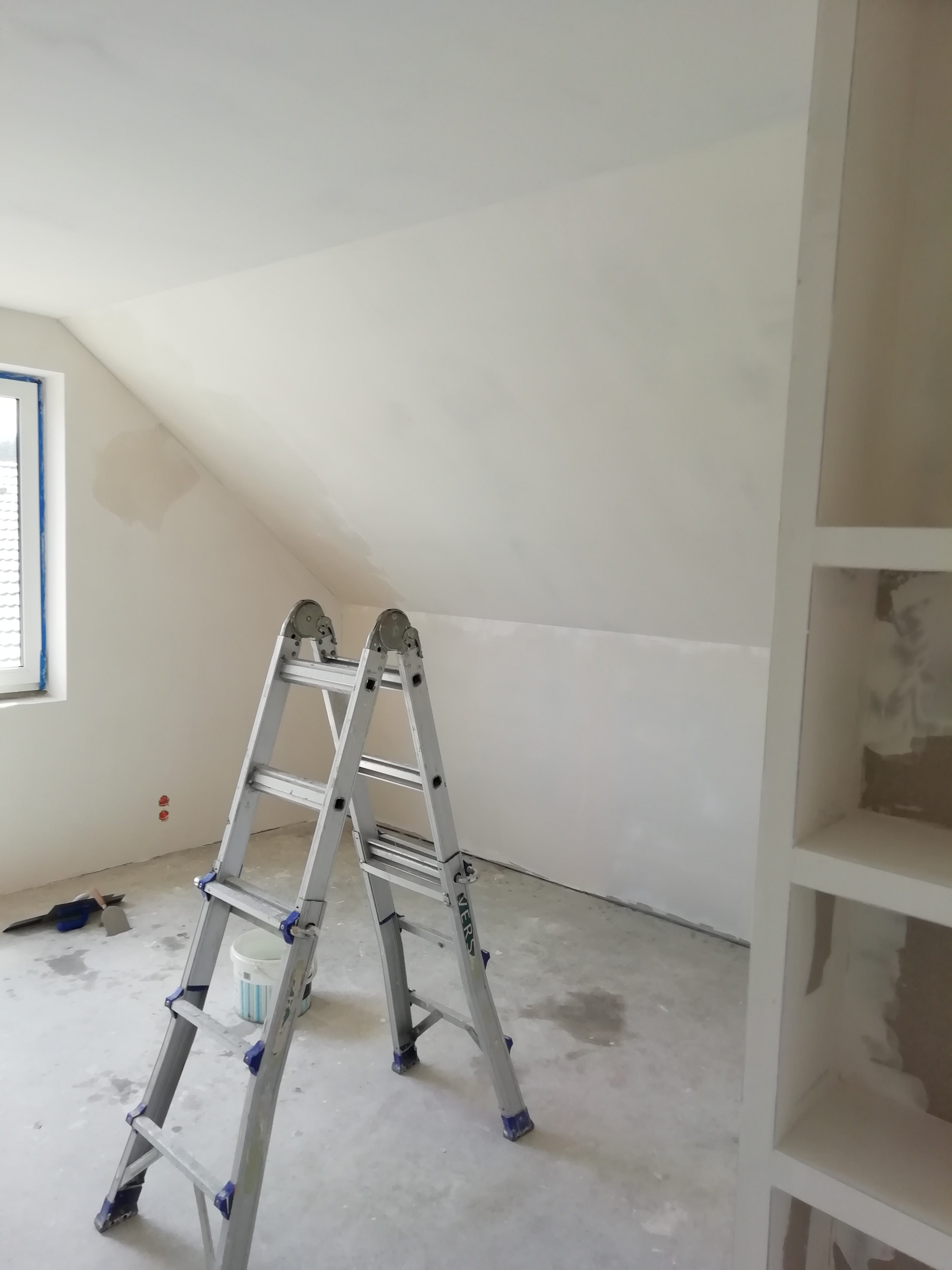 Poddasze w trakcie gładzenia ścian, widoczna aluminiowa drabina, wiadro z gładzią, szpachelki, zaklejone okno i regał wnękowy z resztkami gładzi w środku.