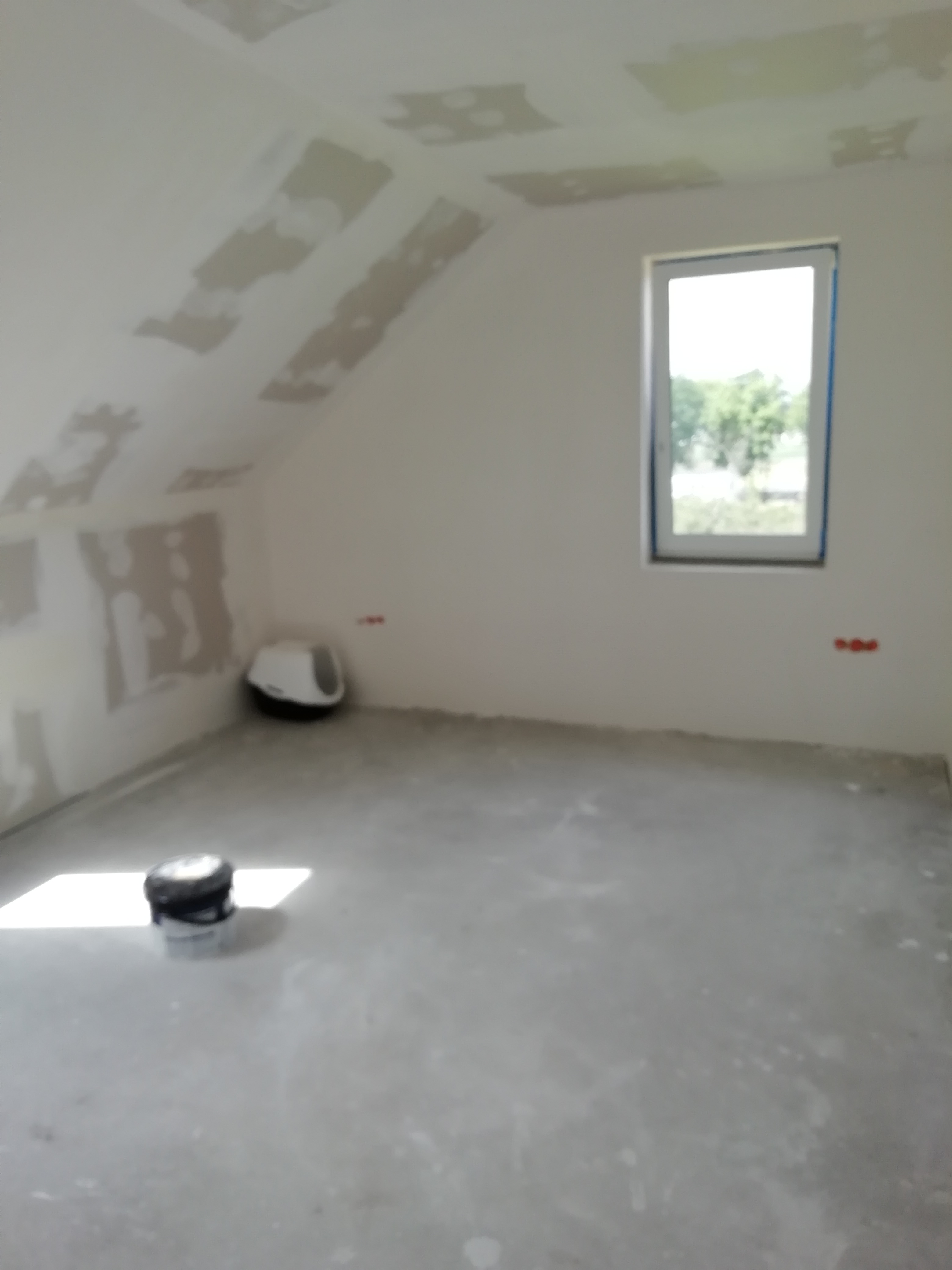 Pokój na poddaszu w trakcie prac wykończeniowych; widoczne fragmenty płyt kartonowo-gipsowych na skosach sufitu przed szpachlowaniem, podłoga przygotowana pod wylewkę, narzędzia i pojemnik z masą...