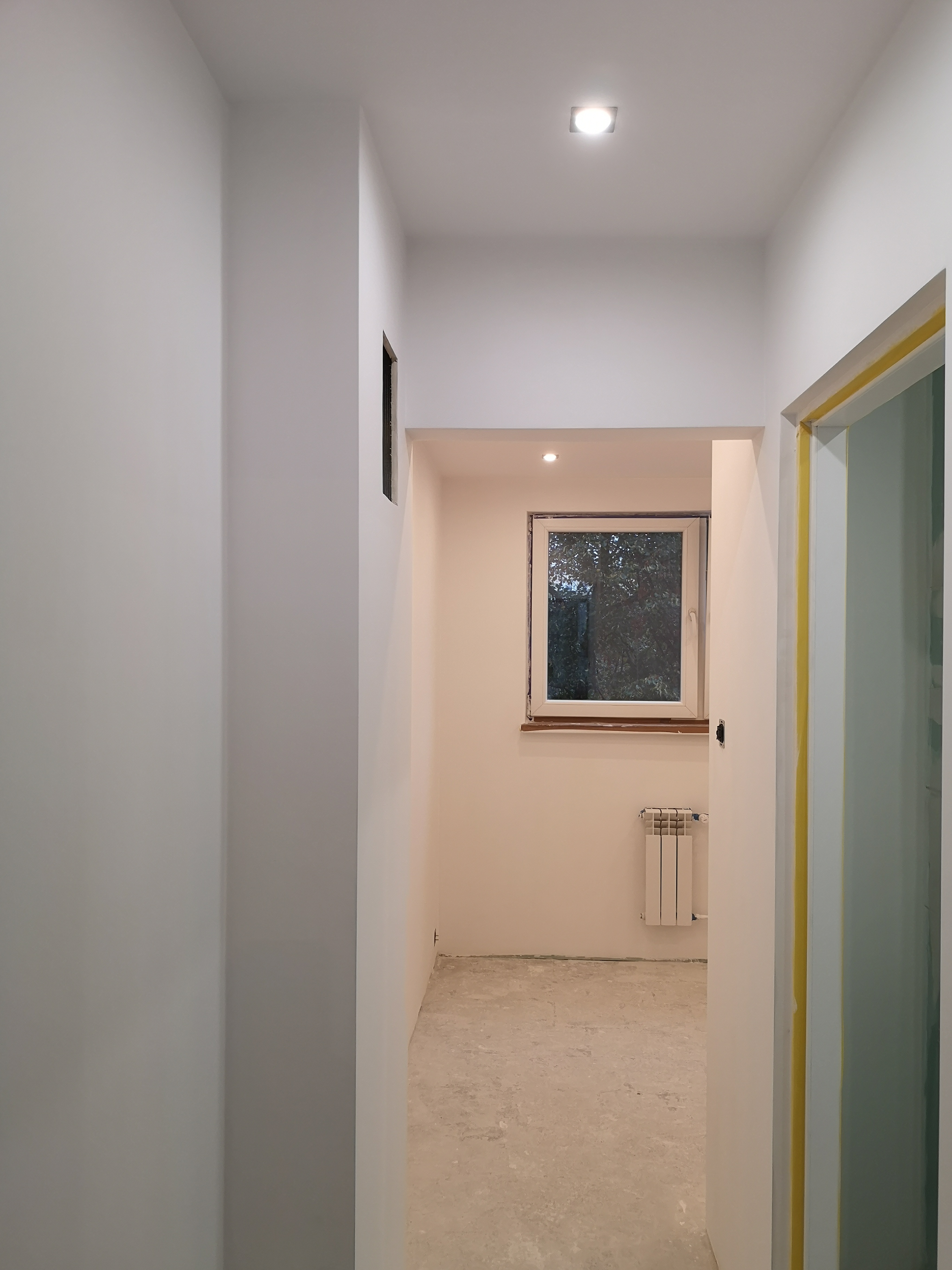 Wnętrze w trakcie malowania na biało, widoczny fragment korytarza z oknem, grzejnikiem i fragmentem futryny zabezpieczonej żółtą taśmą malarską.