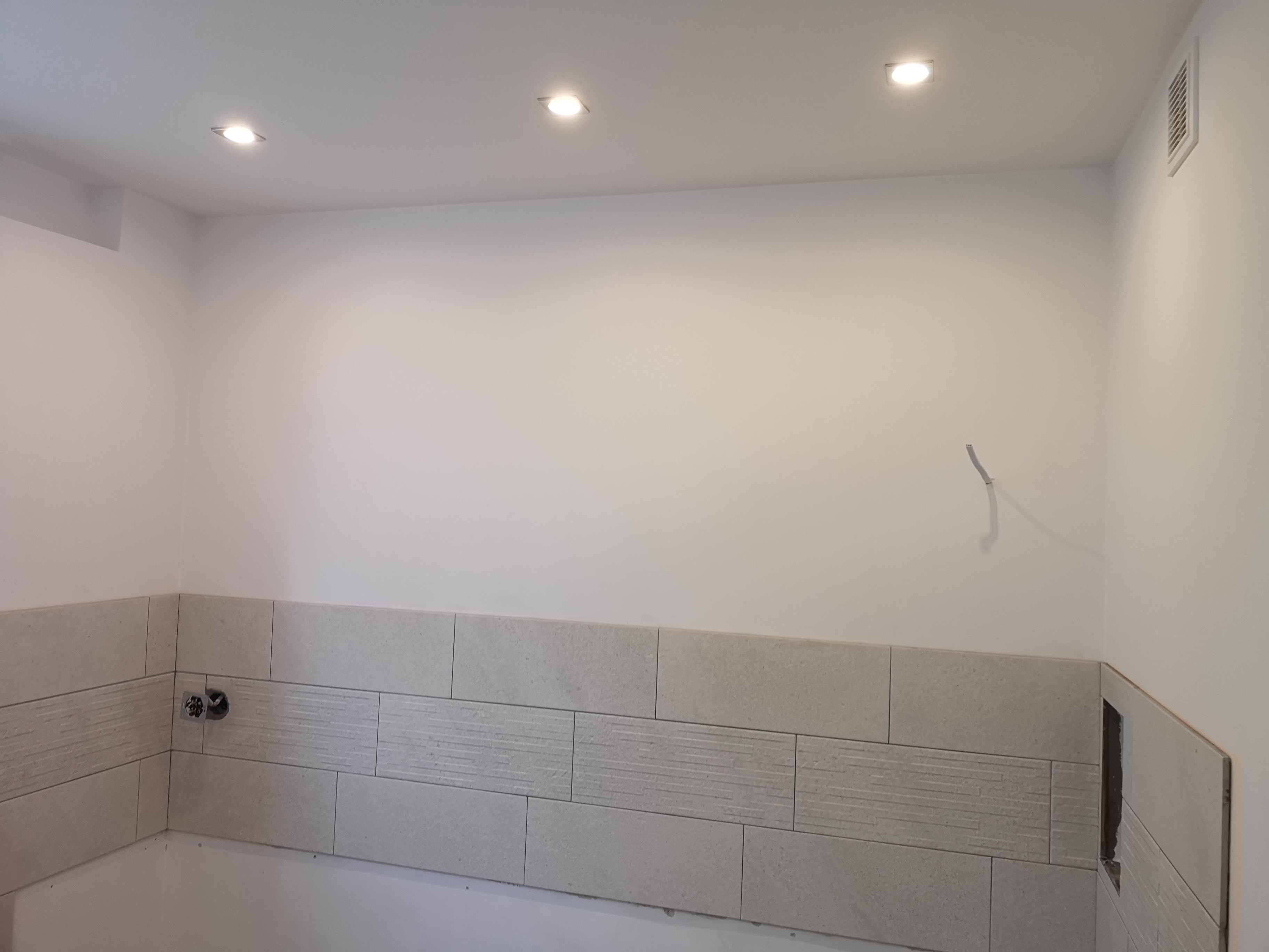 Ściana łazienki w trakcie wykańczania, widoczne ułożone płytki ceramiczne z poziomym wzorem i otwór na gniazdko elektryczne, częściowo ułożone płytki na ścianie bocznej, wystający kabel ze ściany...