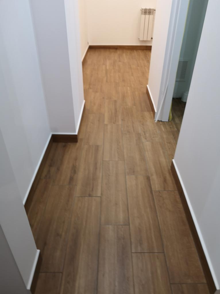 Korytarz z nową podłogą z płytek imitujących drewno, widoczne białe ściany i brązowe listwy przypodłogowe.