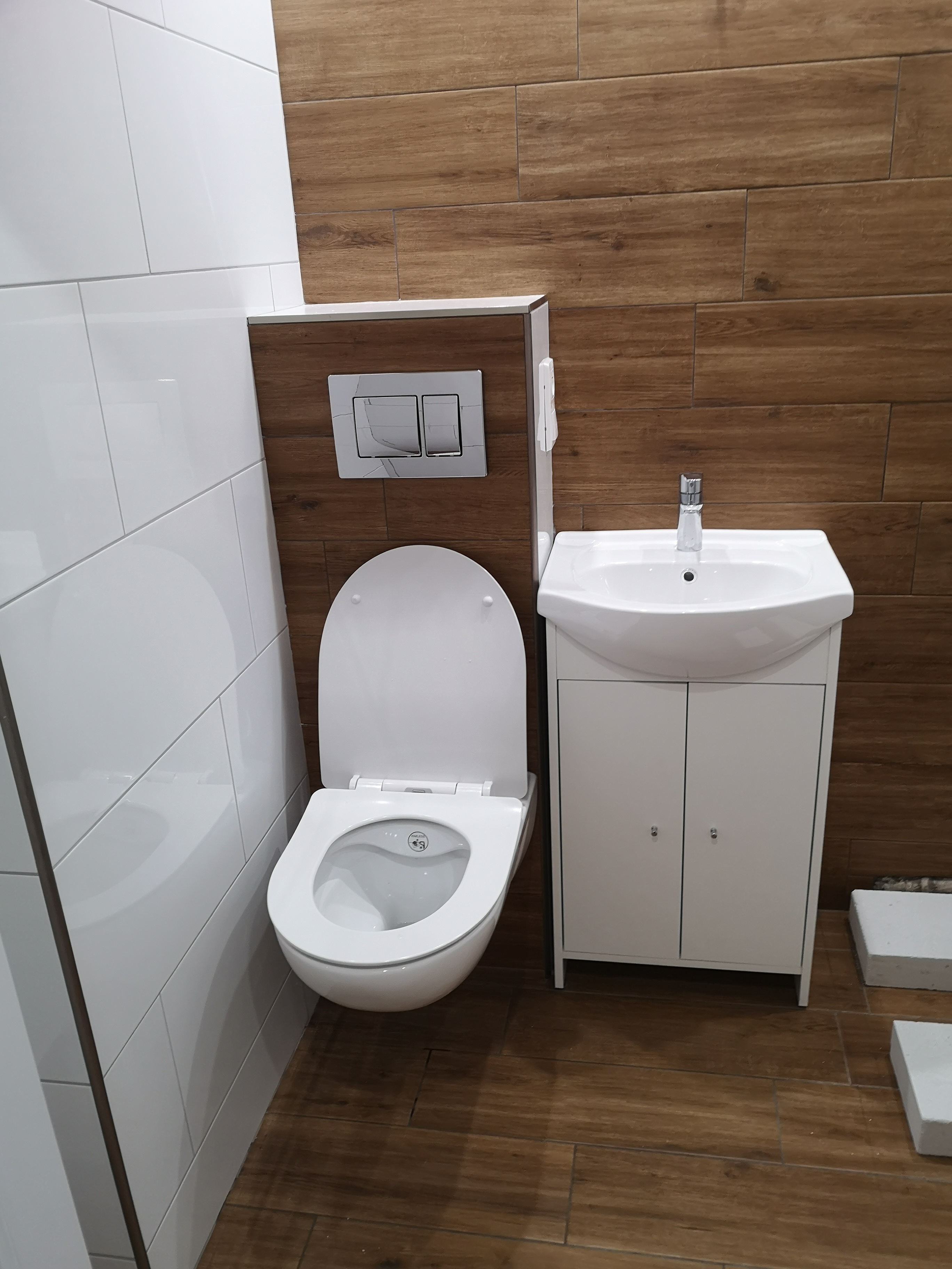 Nowoczesna łazienka z białymi i drewnopodobnymi płytkami, podwieszana toaleta z przyciskiem spłukującym o marmurkowym wzorze, biała umywalka z szafką, widoczne białe bloczki na podłodze.