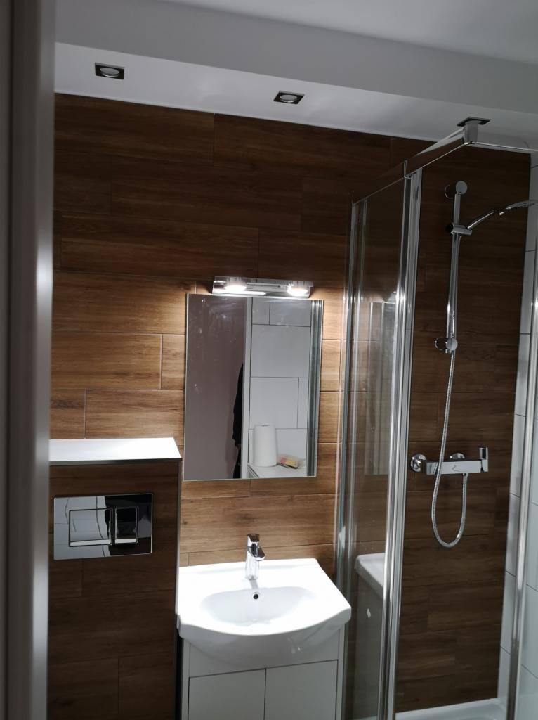 Nowoczesna łazienka z brązowymi płytkami imitującymi drewno, kabiną prysznicową z chromowanymi elementami, umywalką na szafce i lustrem z oświetleniem, widoczne wc podtynkowe.