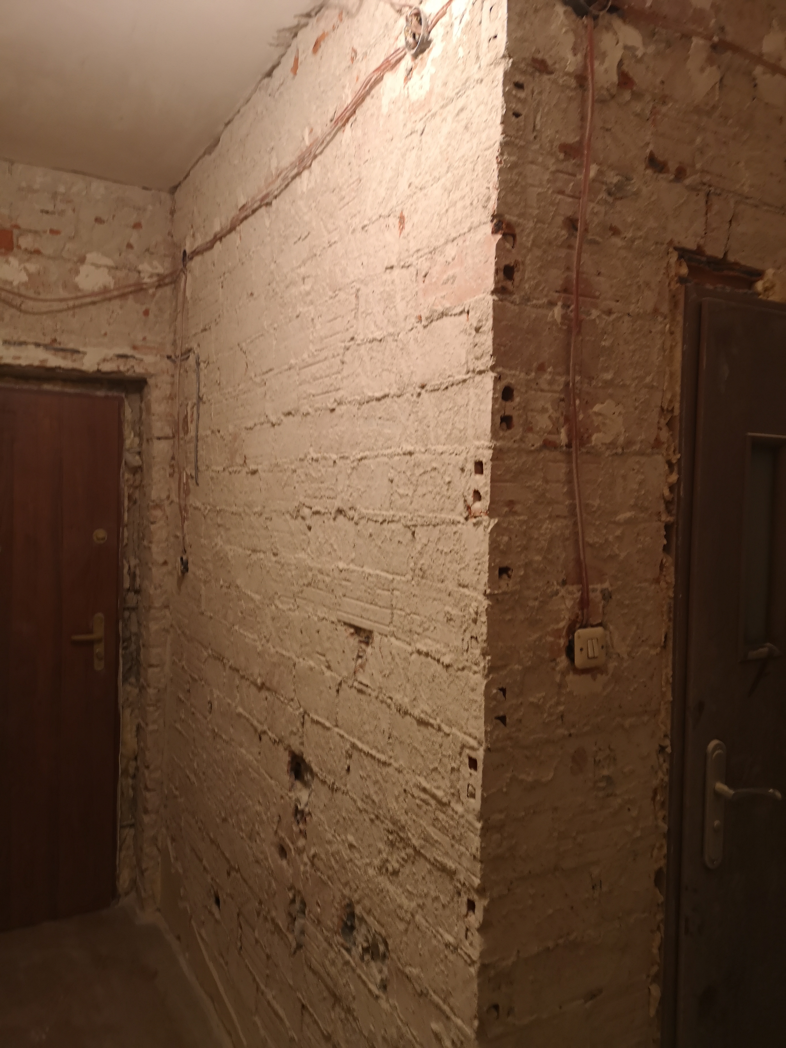 Ceglana ściana w trakcie remontu, częściowo pokryta gładzią, widoczne przewody elektryczne i włącznik światła.