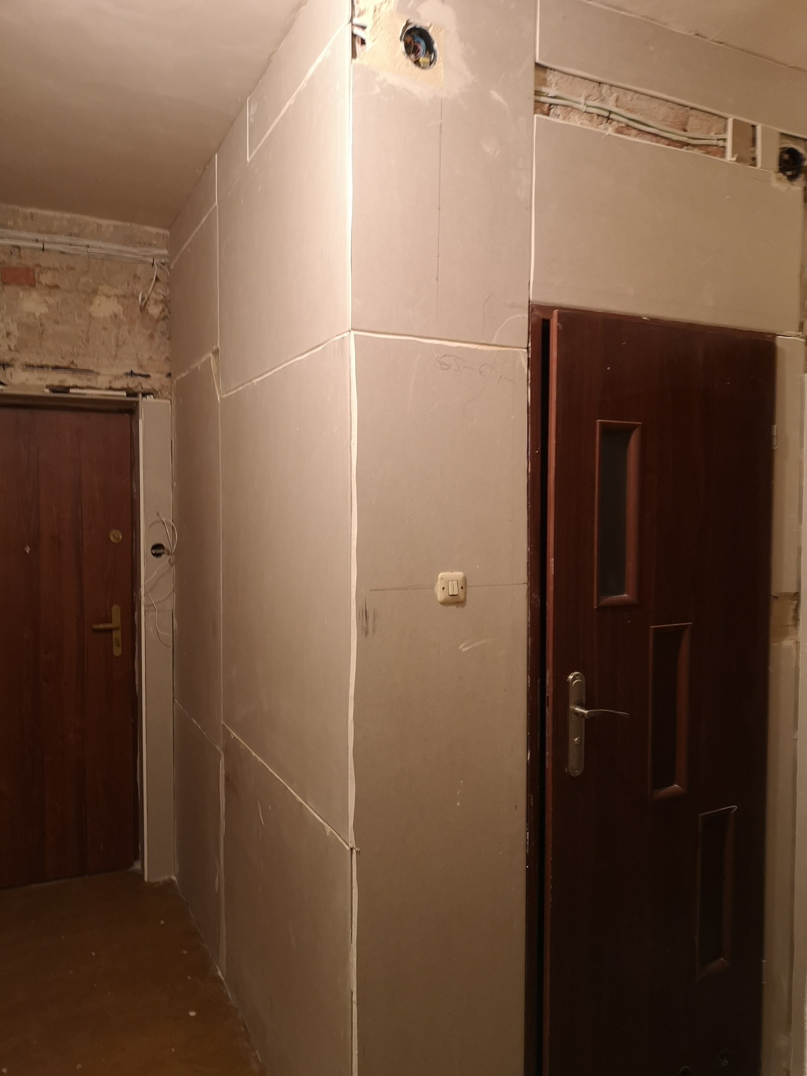 Korytarz w trakcie prac wykończeniowych: zamontowane płyty kartonowo-gipsowe na ścianach, widoczne kable elektryczne, surowa ściana z cegły w tle, brązowe drzwi z ościeżnicą.