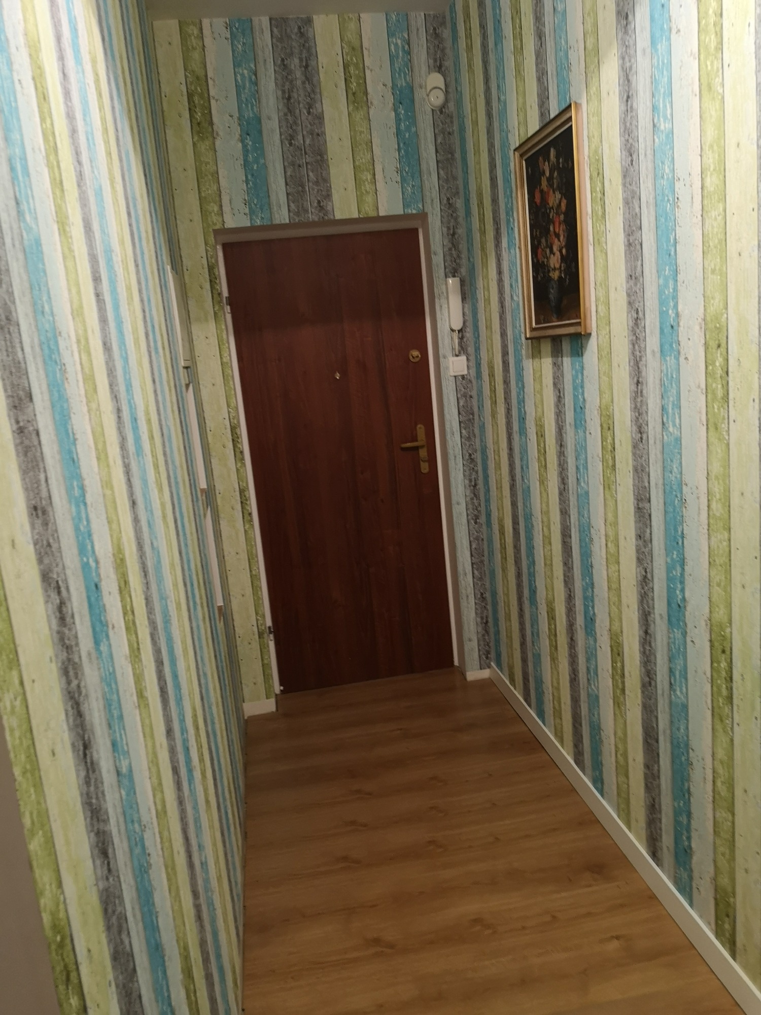 Korytarz po remoncie z drewnianą podłogą, pionowymi tapetami w odcieniach zieleni, błękitu i szarości, brązowymi drzwiami wejściowymi i obrazem w złotej ramie.