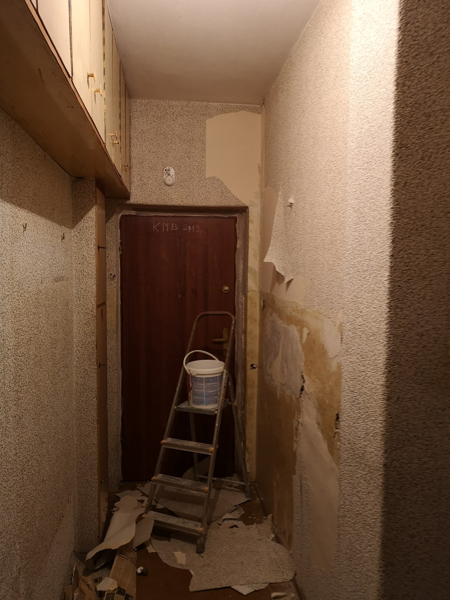 Korytarz w trakcie usuwania starej tapety, widoczna drabina z wiadrem farby i fragmenty oderwanej tapety na podłodze.
