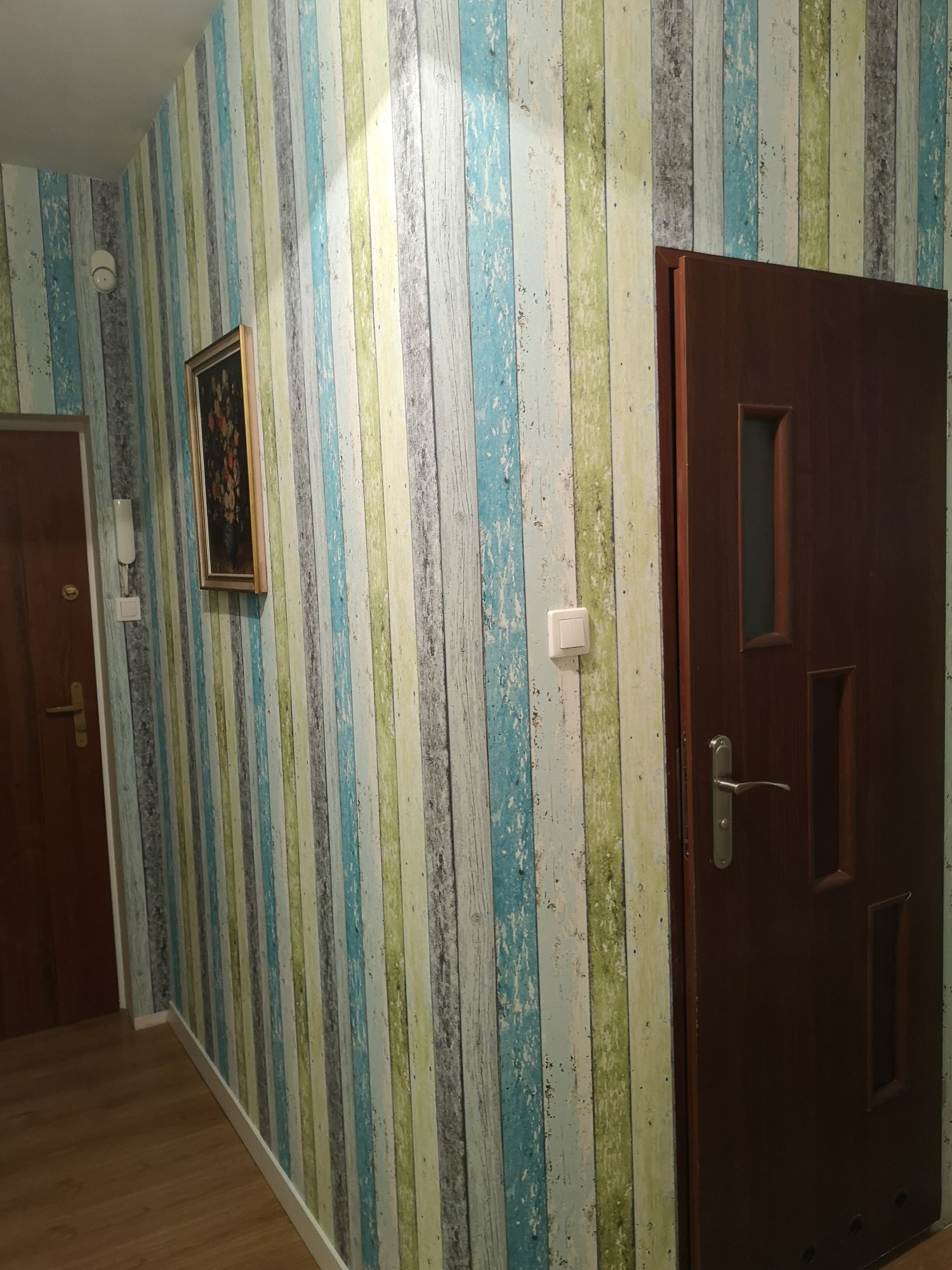 Wnętrze korytarza z drewnianą okleiną na ścianach w odcieniach niebieskiego, zielonego i szarego, obraz w złotej ramie, brązowe drzwi z metalową klamką i widocznym włącznikiem światła.