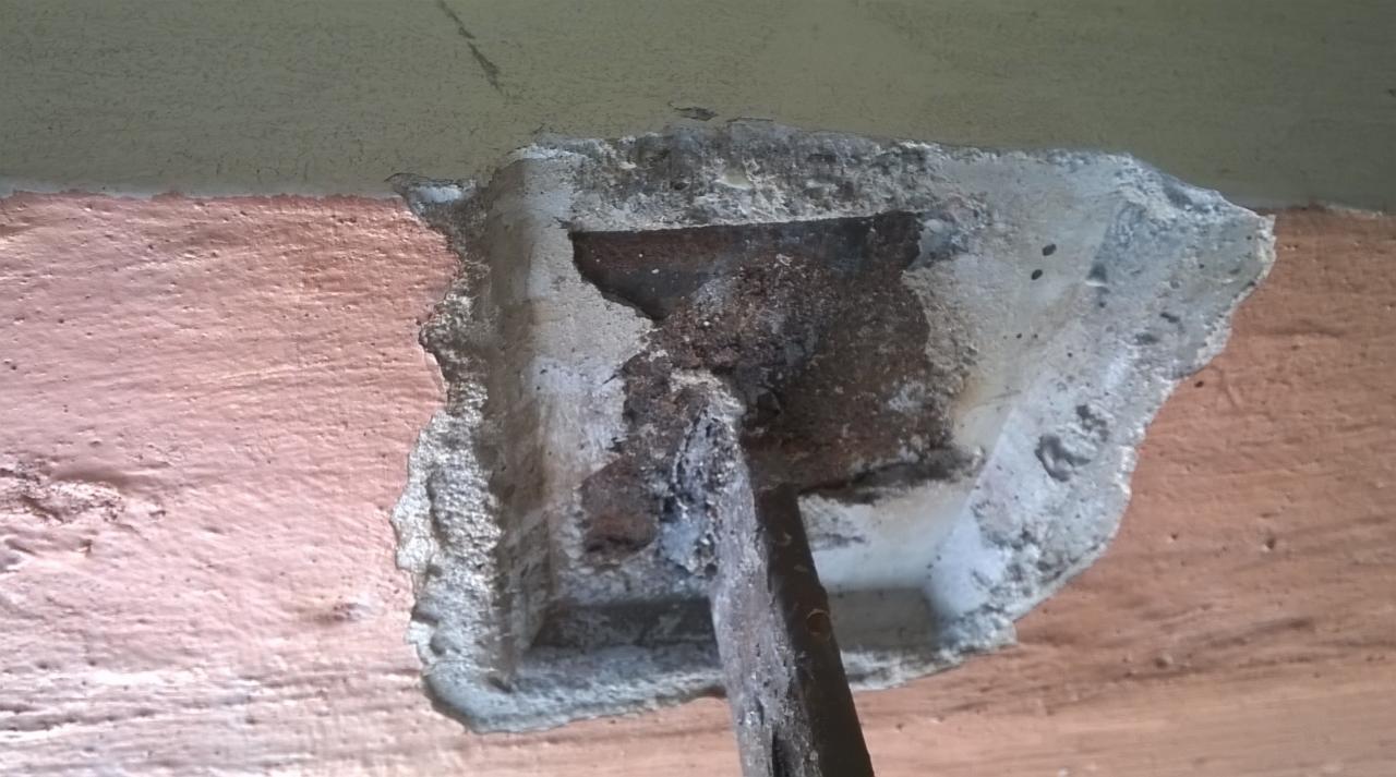 Fragment ściany z uszkodzonym tynkiem, widoczna metalowa szpachelka w otworze, obok różowa pomalowana powierzchnia i fragment sufitu.