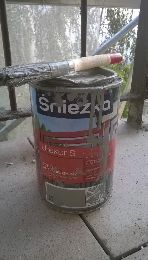 Używana puszka farby podkładowej Śnieżka Urekor S z pędzlem na pokrywie, widoczne zaschnięte ślady farby, tło stanowią rusztowania i zieleń.