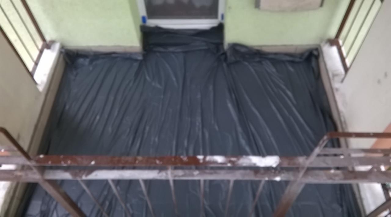 Balkon zabezpieczony czarną folią przed rozpoczęciem prac remontowych, widok z góry z metalowej balustrady.