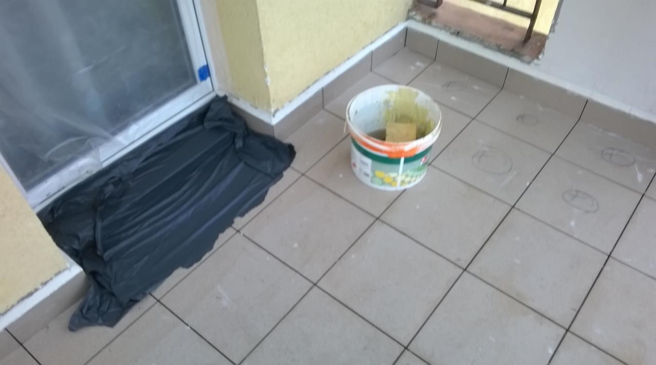 Przygotowania do prac wykończeniowych na balkonie: okno zabezpieczone folią, wiadro z narzędziami i ślady ołówka na płytkach.