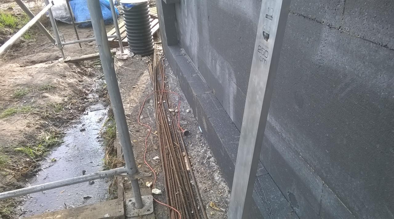 Wyrównana ściana fundamentowa z hydroizolacją i ociepleniem z XPS, widoczny poziomica 250cm, pręty zbrojeniowe oraz rusztowanie w tle.