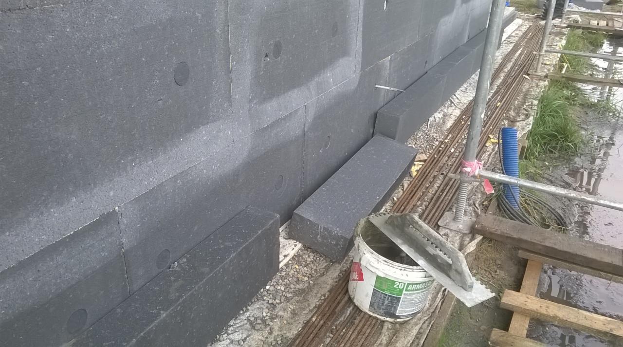 Montaż grafitowego styropianu na ścianie budynku, widoczny kubeł z klejem i szpachla, teren budowy z elementami rusztowania.