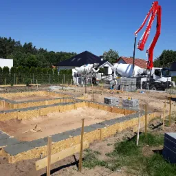 Wylewanie fundamentów pod dom jednorodzinny z użyciem szalunków i pompy do betonu na tle zieleni i zabudowy jednorodzinnej.