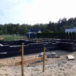 Fundamenty domu jednorodzinnego w Krzeszycach, wykonane z bloczków betonowych, zabezpieczone czarną izolacją, widoczne szalunki i przygotowane materiały budowlane na piaszczystym podłożu.