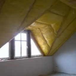 Poddasze z oknem trójkątnym, ocieplone żółtą wełną mineralną pokrytą folią, widoczna konstrukcja drewniana dachu, ściany przygotowane do wykończenia.