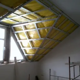 Skośny sufit poddasza w trakcie izolacji, widoczne metalowe profile i żółta wełna mineralna, okno w tle.