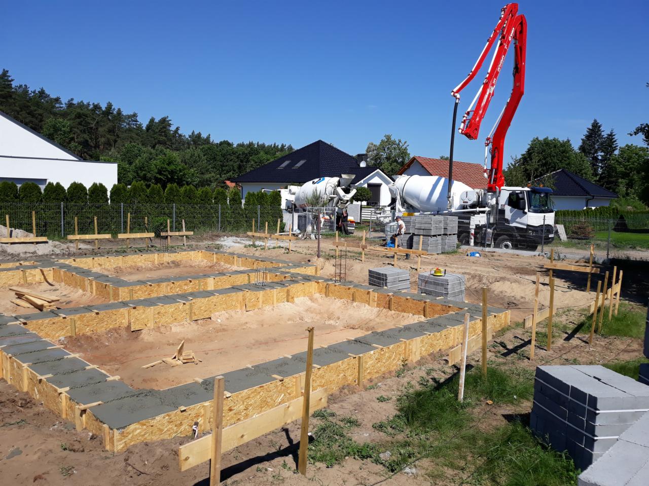 Wylewanie fundamentów pod dom jednorodzinny z użyciem szalunków i pompy do betonu na tle zieleni i zabudowy jednorodzinnej.