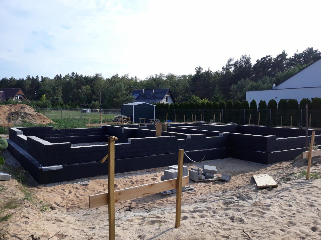 Fundamenty domu jednorodzinnego w Krzeszycach, wykonane z bloczków betonowych, zabezpieczone czarną izolacją, widoczne szalunki i przygotowane materiały budowlane na piaszczystym podłożu.