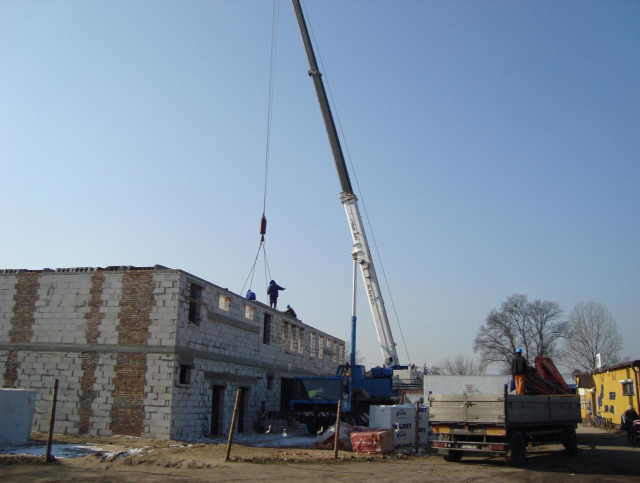 Wznoszenie dwukondygnacyjnego budynku z betonu komórkowego na placu budowy; robotnicy w niebieskich kombinezonach pracują na dachu, a dźwig podnosi materiały budowlane; w tle widoczny samochód...