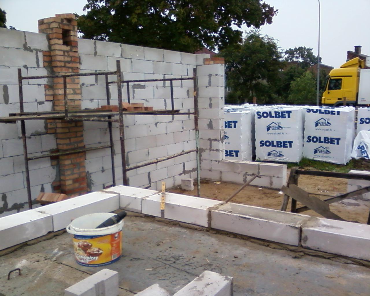 Wznoszenie ścian z białych bloczków Solbet, widoczny fragment komina z cegły oraz rusztowanie, w tle zapakowane bloczki i żółta ciężarówka.