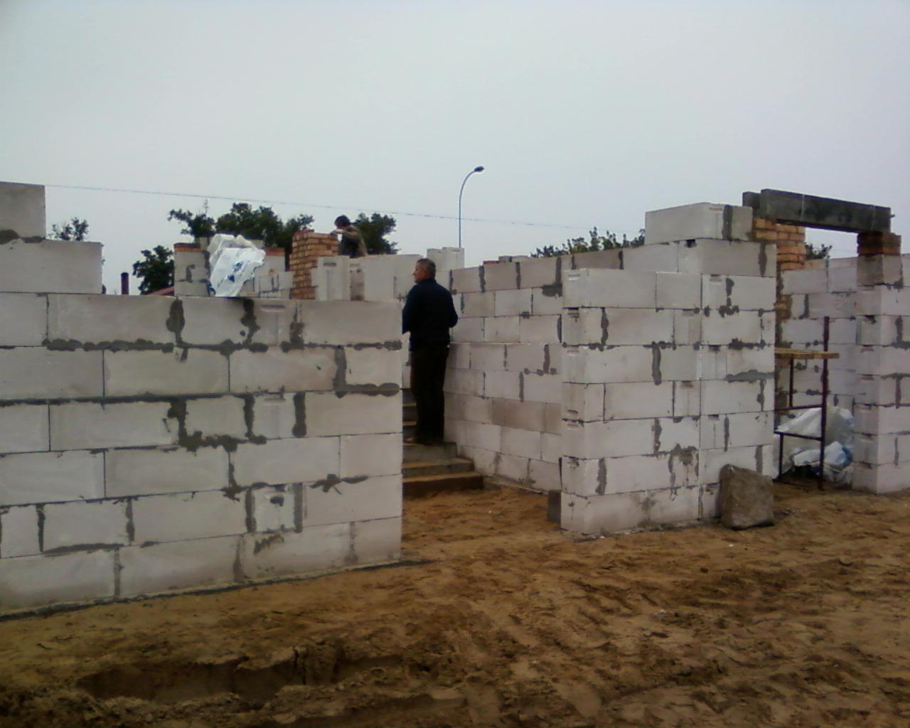 Wznoszenie ścian zewnętrznych domu z betonu komórkowego; widoczny murarz w ciemnym ubraniu na schodach, ściany w trakcie budowy z widocznymi spoinami i zaprawą, teren budowy z piaskiem.