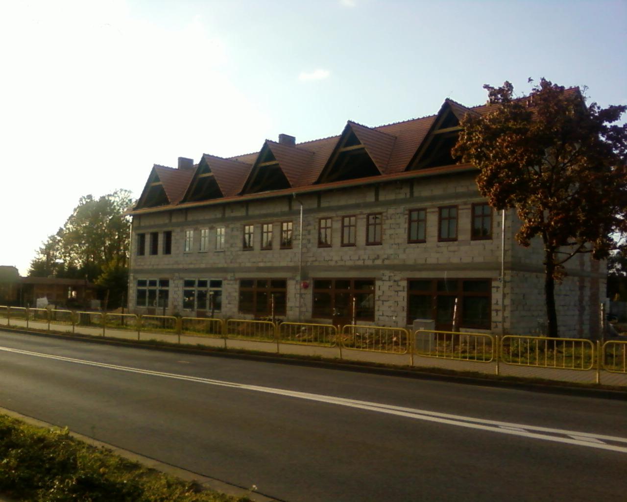 Budynek w stanie surowym zamkniętym z szarego pustaka, z ciemnymi oknami i dachem z czerwonej dachówki, widok z ulicy w słoneczny dzień.