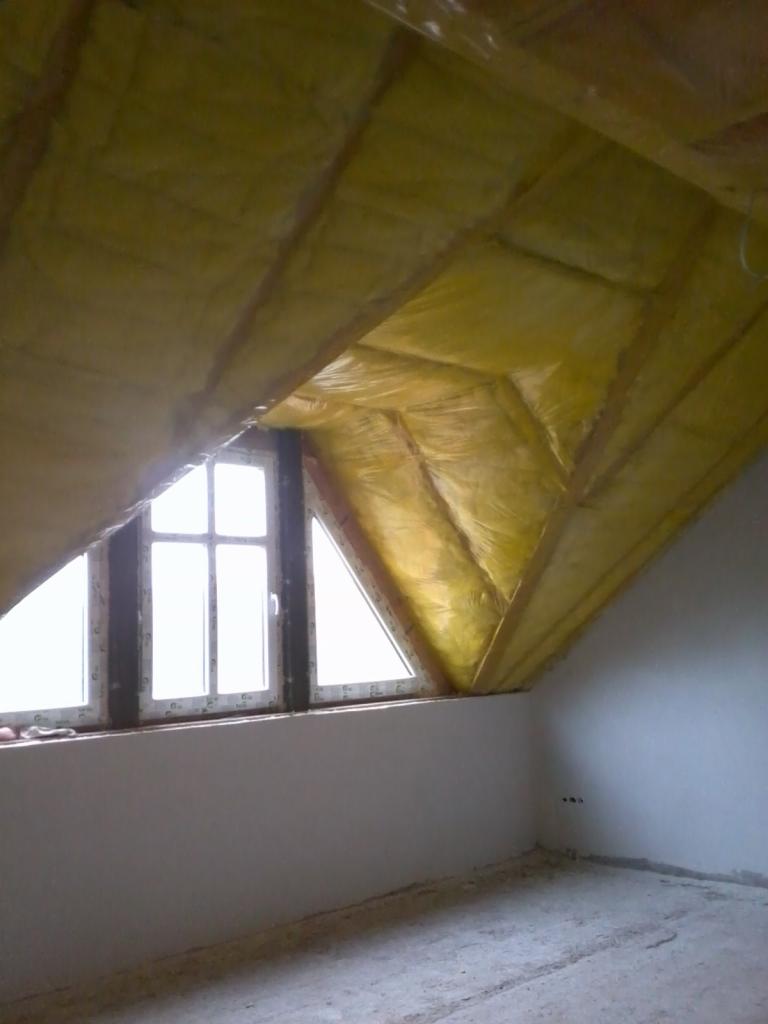Poddasze z oknem trójkątnym, ocieplone żółtą wełną mineralną pokrytą folią, widoczna konstrukcja drewniana dachu, ściany przygotowane do wykończenia.
