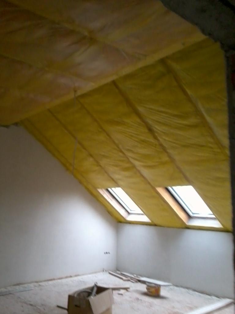 Poddasze w trakcie izolacji, widoczne żółte maty izolacyjne między krokwiami dachu i okna dachowe, surowe ściany i podłoga w nieukończonym pomieszczeniu.