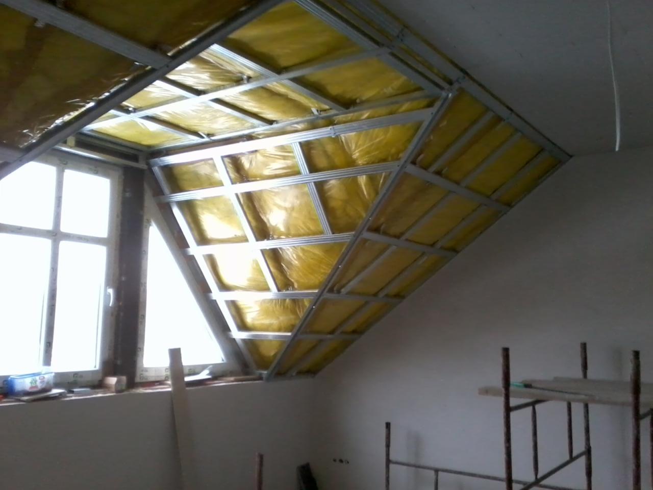 Skośny sufit poddasza w trakcie izolacji, widoczne metalowe profile i żółta wełna mineralna, okno w tle.
