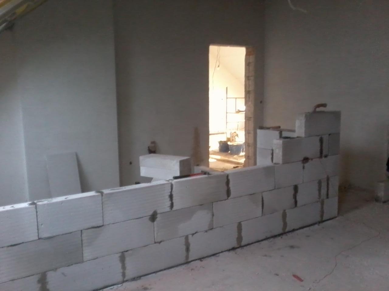 Wznoszenie ściany działowej z białych bloczków na poddaszu, widoczne narzędzia murarskie i fragment otworu drzwiowego z widokiem na strych.