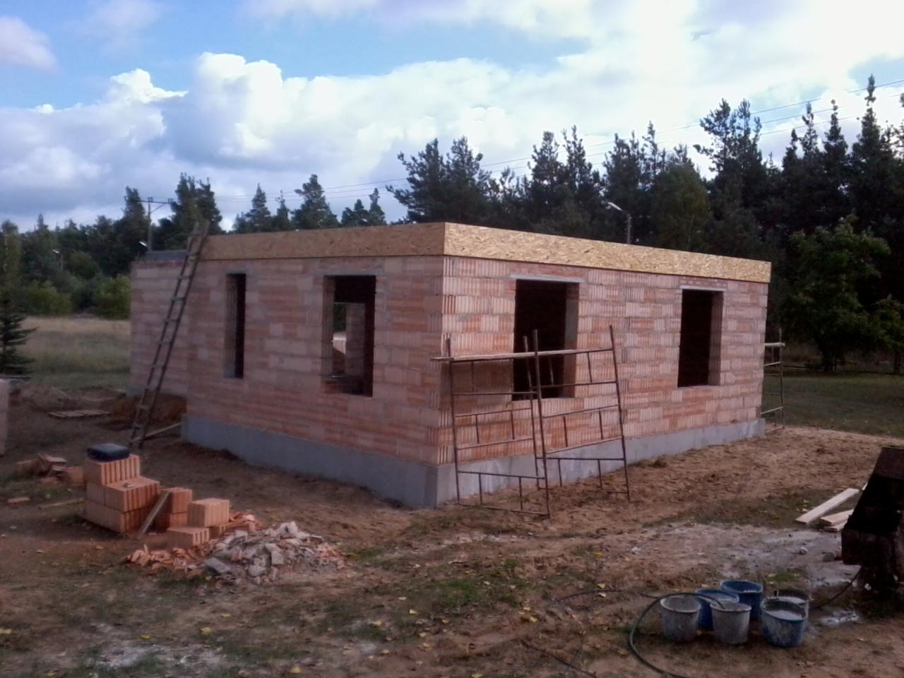 Niedokończony budynek z czerwonej cegły z widocznymi otworami okiennymi, drewnianym szalunkiem na dachu i stosem cegieł obok