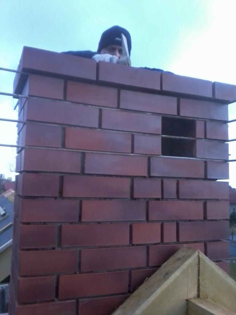 Murarz w czapce i rękawicach buduje komin z czerwonej cegły, widoczny fragment konstrukcji drewnianej.