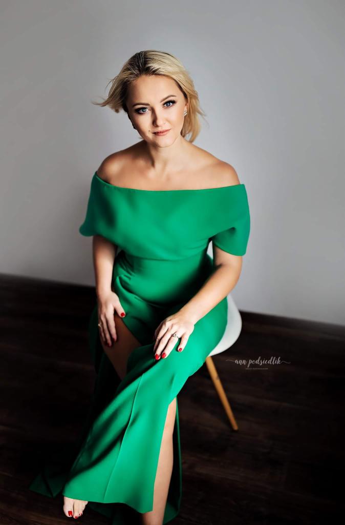 Portret kobiety w szmaragdowej sukni z odkrytymi ramionami, siedzącej na białym krześle. Uśmiech, jasne włosy, subtelny makijaż i czerwone paznokcie.