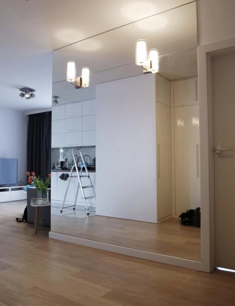 Duże lustro ścienne w nowoczesnym salonie, odbijające kuchnię z drabinką, jasne oświetlenie sufitowe i drewnianą podłogę.