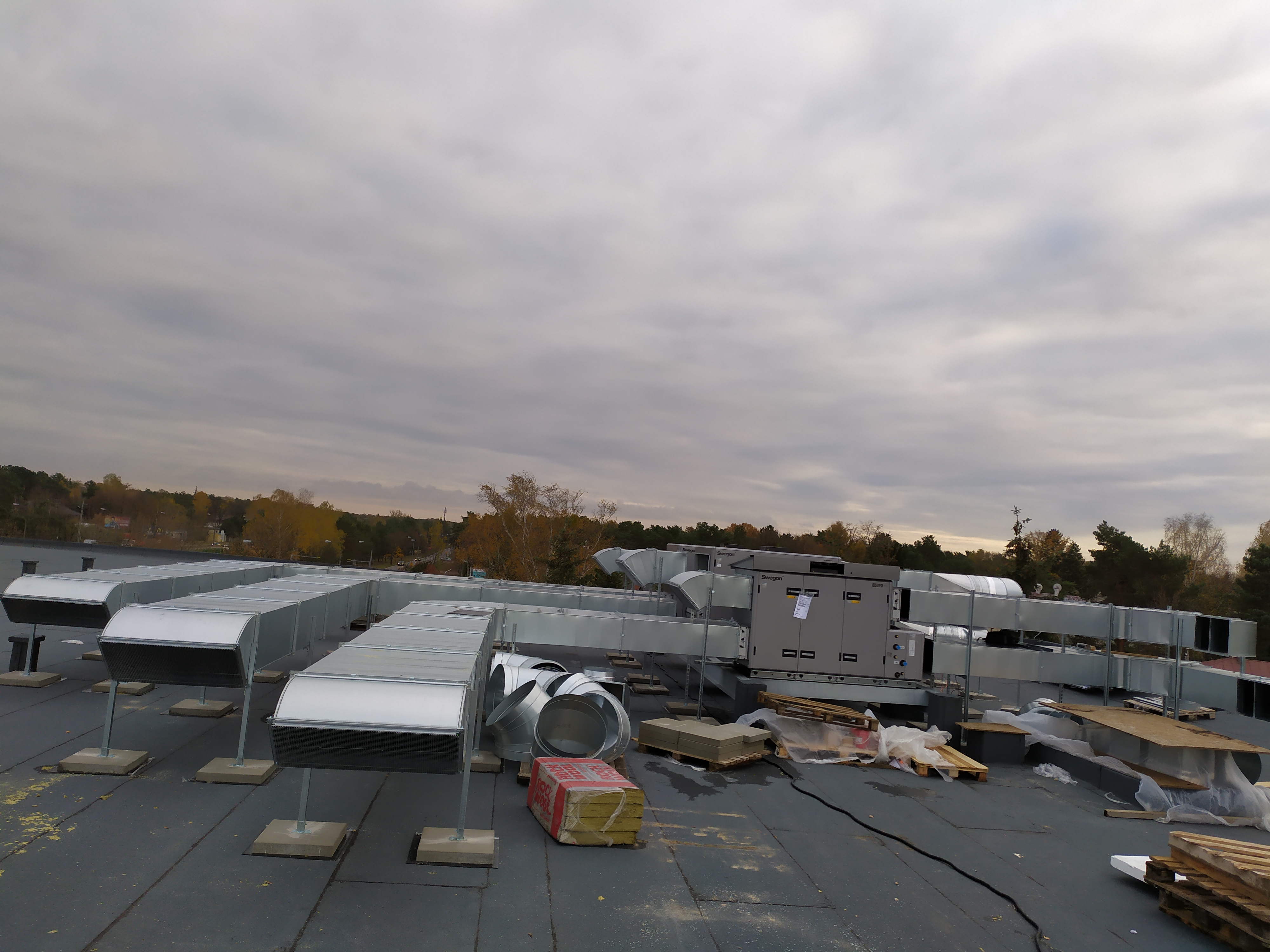 Widok z góry na system kanałów wentylacyjnych na dachu budynku w Mniowie, z widocznymi elementami montażowymi i urządzeniem wentylacyjnym marki Swegon, na tle pochmurnego nieba i jesiennego...