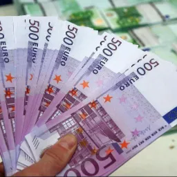 Dłoń trzyma wachlarz banknotów 500 euro na tle stosów banknotów.