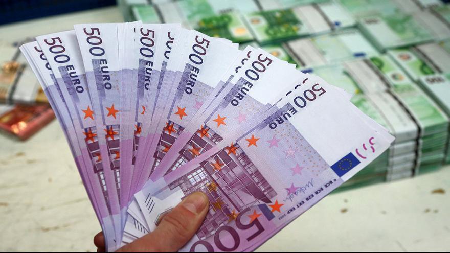Dłoń trzyma wachlarz banknotów 500 euro na tle stosów banknotów.