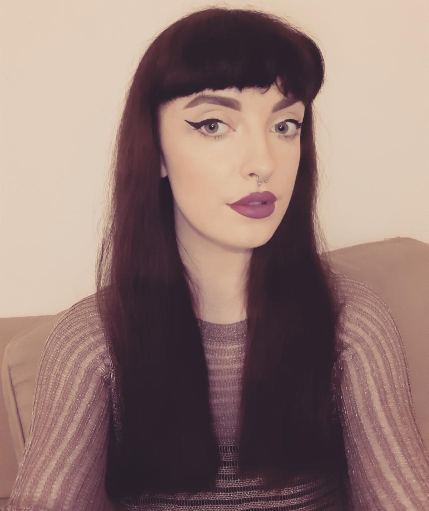 Portret kobiety z ciemnymi włosami, mocnym makijażem ust w odcieniu fuksji i wyraźną kreską na powiekach. Widoczny piercing w nosie i bluzka w paski.